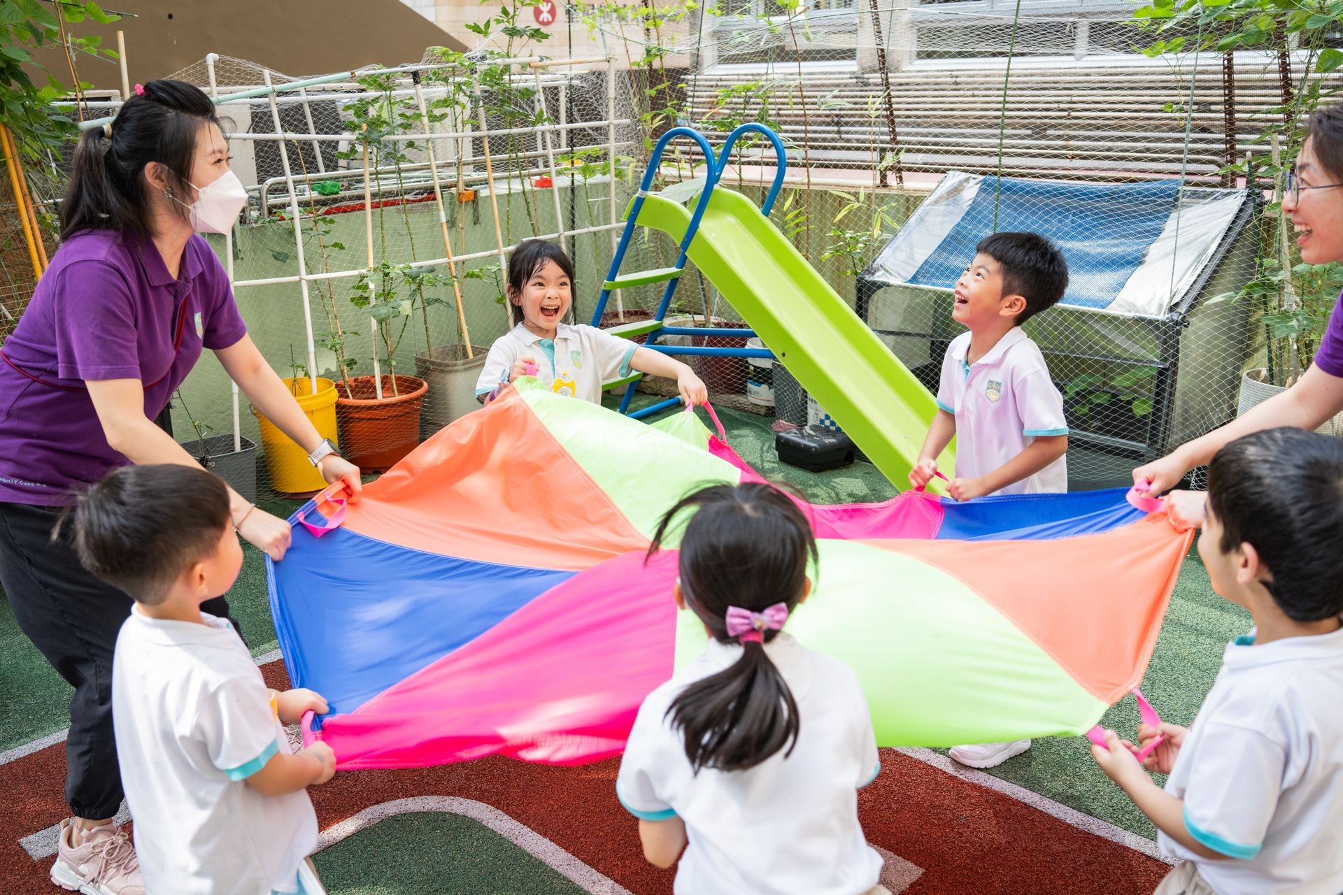 Teacher playing parachute games with laughing children — a joyful moment at our nurturing preschool in Hong Kong